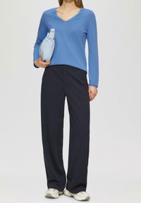 Niebieski sweter z długim rękawem i dekoltem w serek, zestawiony z ciemnymi, szerokimi spodniami. Trzyma jasnoniebieską saszetkę. Buty w kremowym kolorze z siateczkowymi detalami.