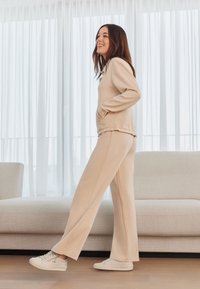 Set in beige composto da felpa e pantaloni in un materiale morbido, con tasche e una vestibilità rilassata, abbinato a sneakers bianche su un pavimento di legno.