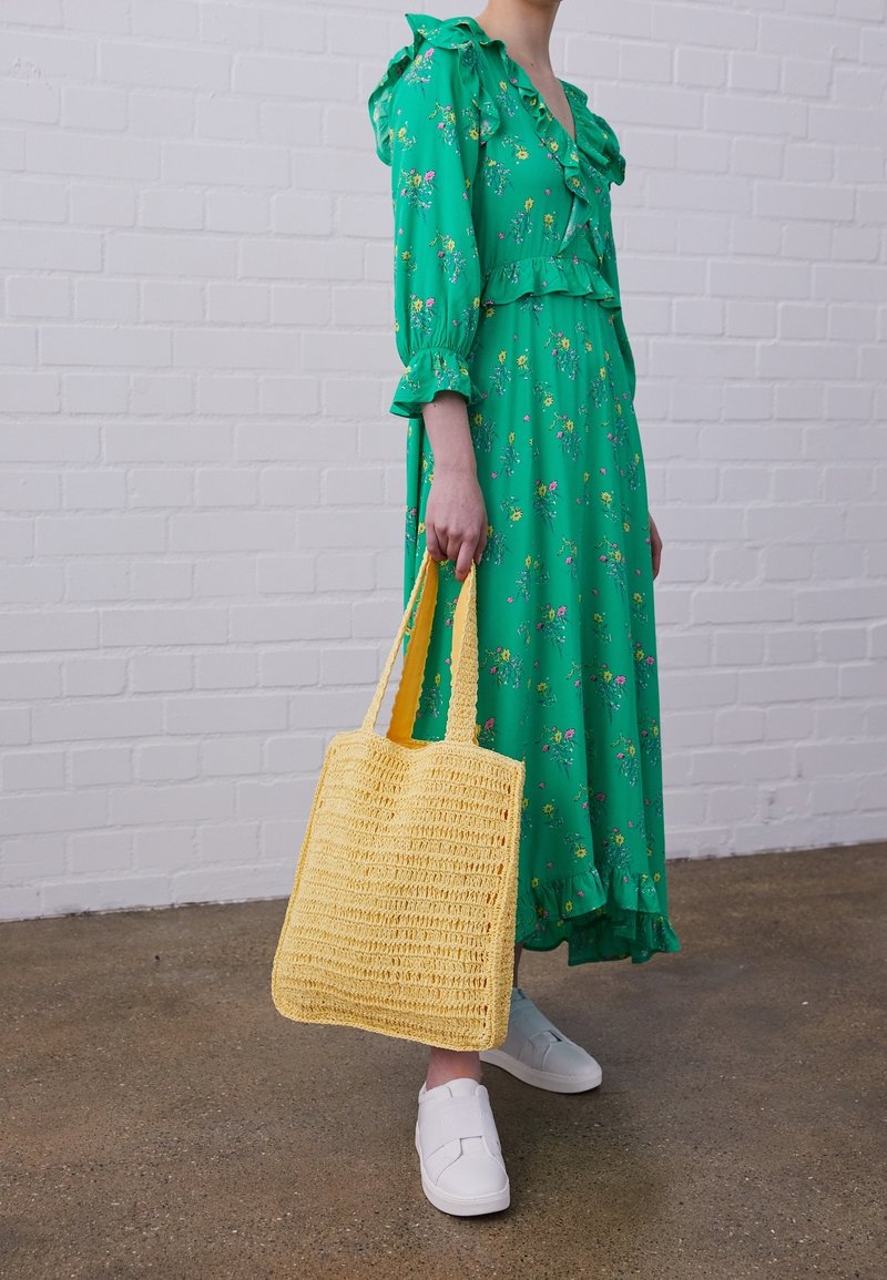 Persoon in een groene floral midi-jurk en witte sneakers, met een grote gele geweven tas, staande op een betonnen vloer.