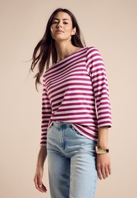 Maglietta a maniche lunghe con audaci righe rosa e bianche, scollo rotondo, abbinata a jeans azzurri e un bracciale a manica d'oro. Tessuto dalla texture liscia.