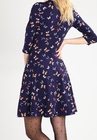 Marinblå klänning med fjärilstryck, som har en åtsittande liv, utsvängd kjol och trekvartsärmar. Bärs med prickiga strumpbyxor.