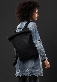 Schwarzer Rolltop-Rucksack mit einer Reißverschlusstasche vorne, gefertigt aus strukturiertem Material. Getragen mit einer abgenutzten Denimjacke. Mattes Finish.