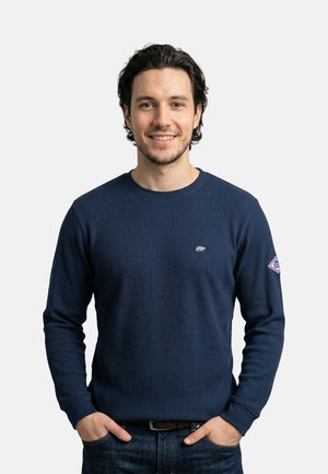 Lachende man met donker krullend haar, gekleed in een navyblauw gestructureerd shirt met lange mouwen en spijkerbroek, handen in de zakken, staand tegen een effen achtergrond.