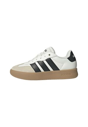 Zapatilla blanca de caña baja con rayas negras, suela color cuero, puntera beige y lengüeta de talón con marca, mostrada desde el lado exterior.