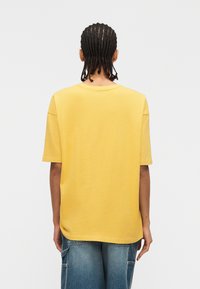 Άτομο με πλεξούδες φοράει ένα φαρδύ κίτρινο κοντομάνικο πουκάμισο και μπλε τζιν παντελόνι cargo, στέκεται μπροστά σε απλό λευκό φόντο, με την πλάτη προς τα εμπρός.