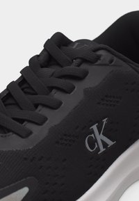 Μαύρο αθλητικό παπούτσι με υφασμάτινη υφή και μαύρα κορδόνια, με λογότυπο "cK" στο πλάι και λευκή σόλα.