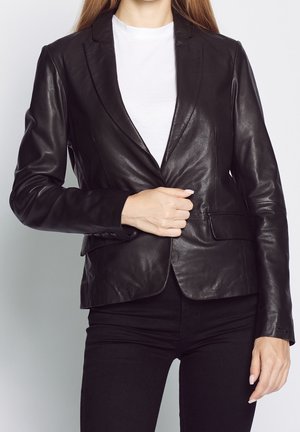 Vrouw draagt een zwarte leren blazer over een wit shirt, terwijl ze de blazer met haar rechterhand dichtknoopt en zwarte broek draagt.