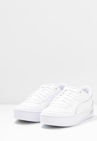 Bílé tenisky s hladkým koženým svrškem, zakulacenou špičkou, plochými tkaničkami a silnou platformou. Obsahují jemné boční logo a detaily stehu.