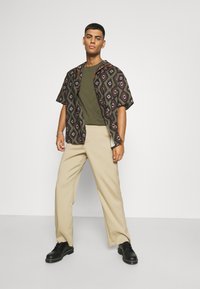 Short-sleeved patterned shirt in dark colors with diamond shapes, olive green t-shirt, light beige trousers, and black shoes.
