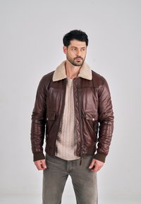 Hnědá kožená bomber bunda s béžovým límcem z beránku, zapínáním na zip a dvěma předními kapsami. Nošená přes texturovaný světlý svetr.