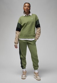 Felpa verde con accenti neri e beige, abbinata a pantaloni jogging verdi coordinati. Presenta polsini a coste e logo sul petto. Sneakers beige.