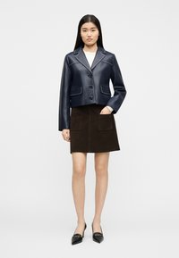 Femme portant une veste en cuir courte bleu marine, un haut blanc, une jupe en daim marron foncé avec des poches frontales, et des talons noirs à bout pointu.