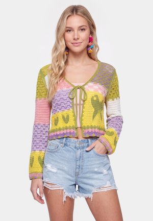 Giovane donna con lunghi capelli biondi indossa un cardigan colorato all'uncinetto su una canottiera rosa e shorts di jeans chiari strappati, sorridendo leggermente.