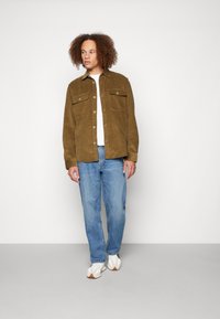 Brun kordfløyelsjakke med to frontlommer, knappelukking, brukt over en hvit T-skjorte, kombinert med lyseblå denim og hvite joggesko.