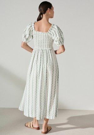 Woman with dark hair in a low ponytail wearing a white, ankle-length dress with puffed sleeves and vertical green floral stripes, standing with hands on hips.