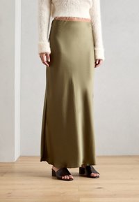 Jupe satin vert olive au design fluide, fente sur le côté et texture lisse ; associée à des sandales à talons ouverts noires.