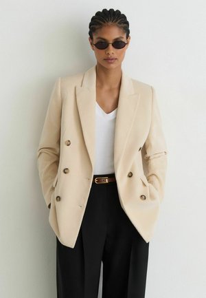 Frau mit geflochtenem Haar, die schwarze ovale Sonnenbrille, einen beigen Doppelreiher-Blazer, ein weißes Top, schwarze Hosen und einen braunen Gürtel trägt und vor einer weißen Wand steht.
