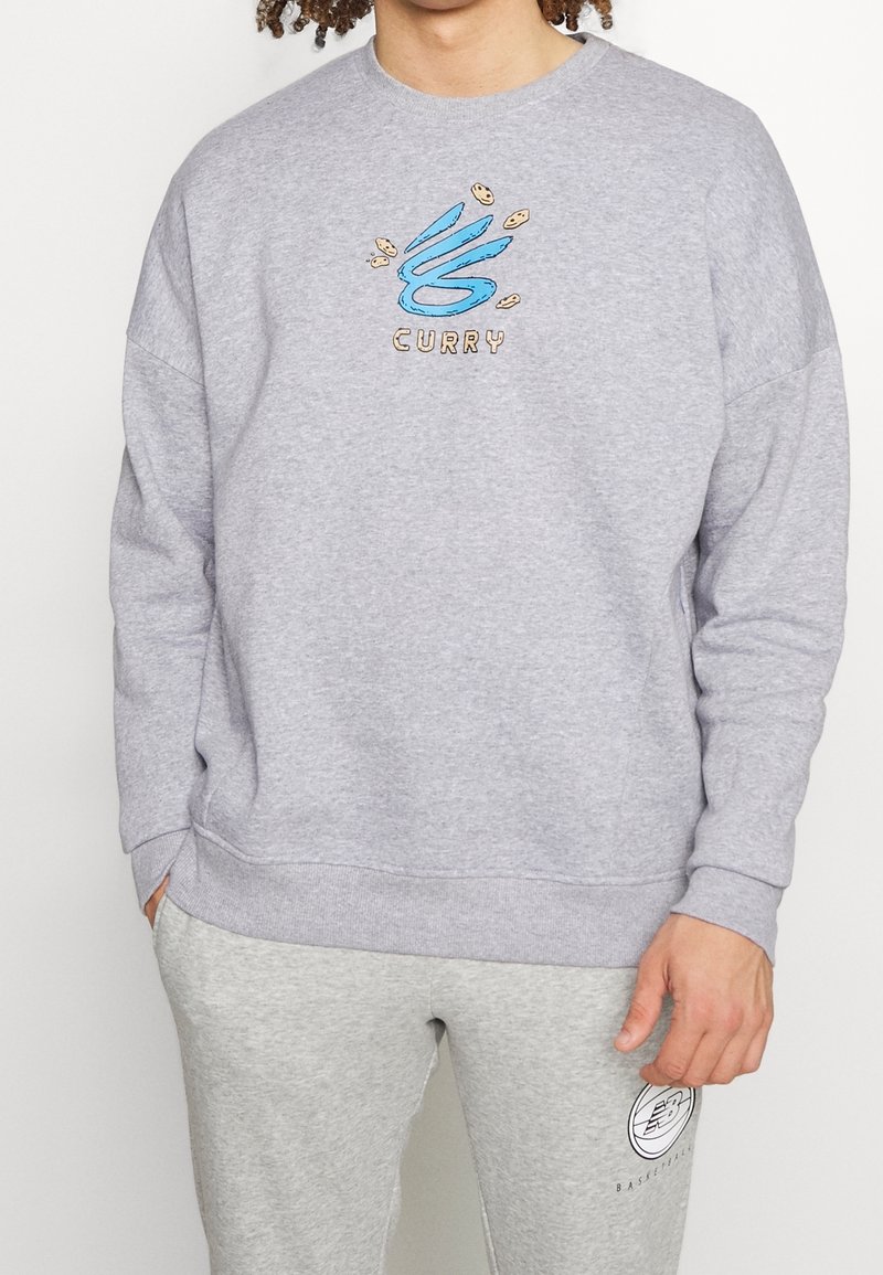 Sweatshirt gris avec un col rond, présentant un design brodé bleu et beige, "CURRY," et une coupe ample. Assorti avec un jogging gris.