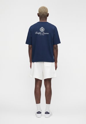 Rövid szőke hajú férfi, aki sötétkék Ralph Lauren pólót, fehér rövidnadrágot, fehér zoknit és fehér tornacipőt visel sötétkék sarokkal, háttal.