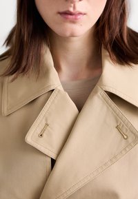 Gros plan d'une personne portant un trench-coat beige avec de larges revers, montrant le col et une partie du visage à partir du nez vers le bas.