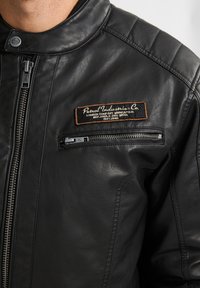 Chaqueta de cuero negra con cuello alzado, cierre delantero con cremallera y un parche etiquetado "Petrol Industries Co." que presenta texto en color marrón claro y blanco.