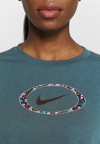 Primo piano di una persona che indossa una maglietta teal con un ovale floreale che circonda un logo Nike nero sul petto.