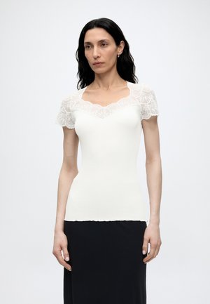 Kvinde med langt sort hår iført en hvid tætsiddende top med blonder korte ærmer og halsudskæring, kombineret med et sort skørt, stående oprejst.