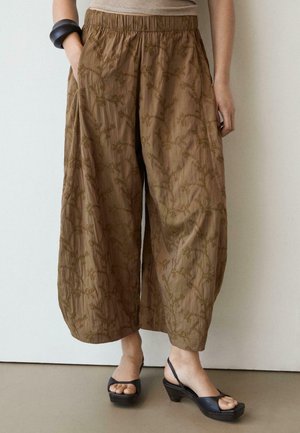 Pantalon marron à jambes larges et courts avec une broderie florale subtile, porté avec des sandales à talons ouvertes noires et un bracelet noir au poignet gauche.