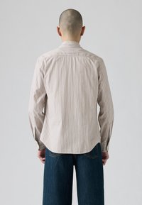 Camicia a maniche lunghe di color beige chiaro con righe verticali in vari colori. Caratterizzata da un colletto piegato con cura e da un orlo curvo.