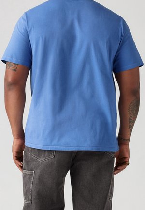 Homme portant une chemise bleue unie à manches courtes et un pantalon cargo gris foncé, vu de dos sur un fond neutre.