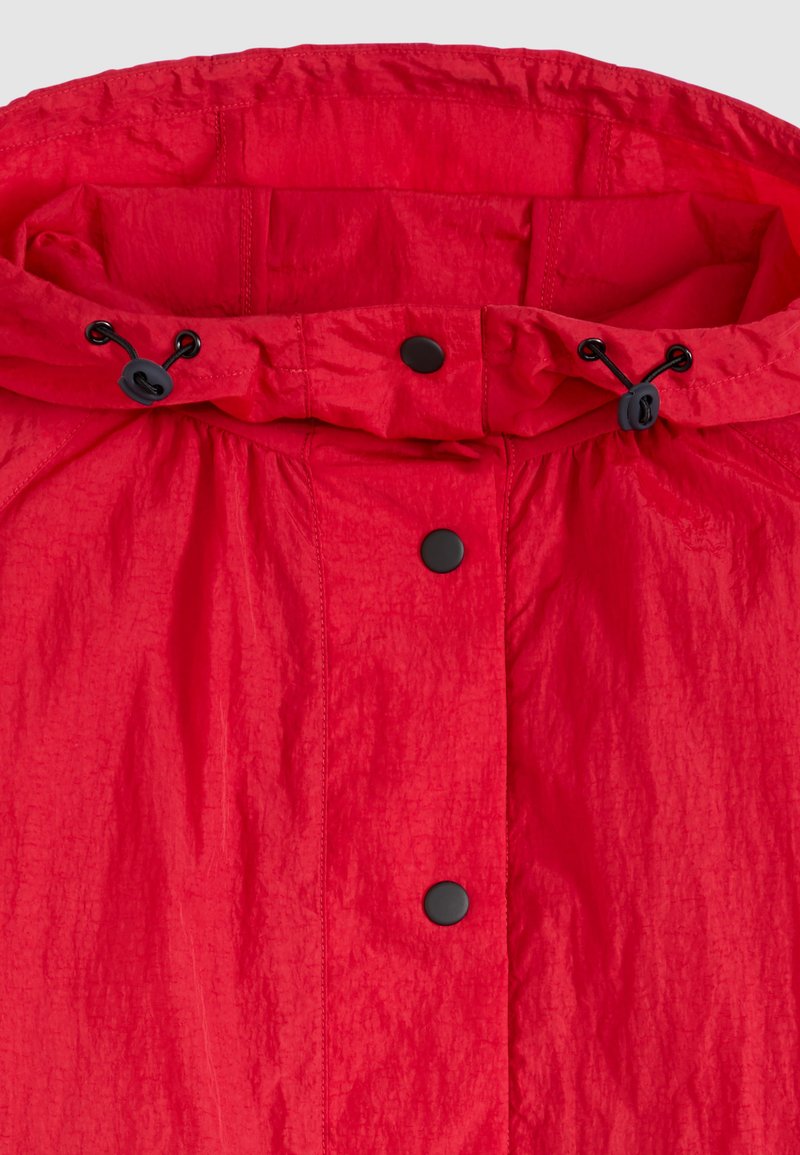 Helle rote Kapuzenjacke mit schwarzen Druckknöpfen und verstellbaren schwarzen elastischen Kordelzügen auf einem strukturierten Stoff.
