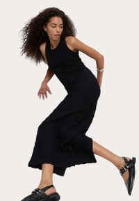 Svart texturerad ärmlös klänning med medellång fåll, i kombination med svarta slingback-skor med metalliska detaljer. Modell i rörelse.