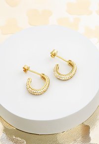 Goldene Creolen mit einem geschwungenen Design, verziert mit klaren, funkelnden Strasssteinen, montiert auf einer weißen runden Präsentationsfläche.