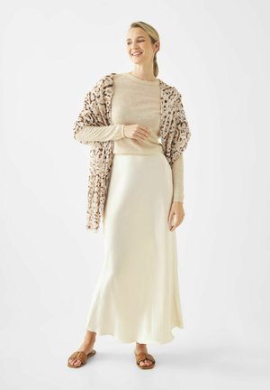 Vrouw glimlachend, gekleed in beige top met lange mouwen, crèmekleurige satijnen rok, sjaal met luipaardprint en bruine sandalen, staand tegen een witte achtergrond.