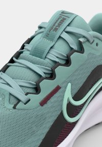 Helle türkisfarbene Sportschuhe aus Netz- und Gummi-Materialien. Merkmale sind eine strukturierte Oberfläche, schwarze und maulbeerfarbene Akzente sowie ein auffälliges Swoosh-Logo.