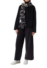 Osoba w czarnej kurtce, czarnych szerokich spodniach, wzorzystym czarno-beżowym szaliku i białych tenisówkach na białym tle.