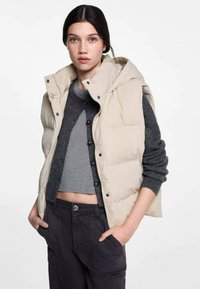 Beige puffer-väst med huva och tryckknapps-stängning, bärs över en mörkgrå croppad tröja, ihop med mörka byxor, har en avslappnad passform.