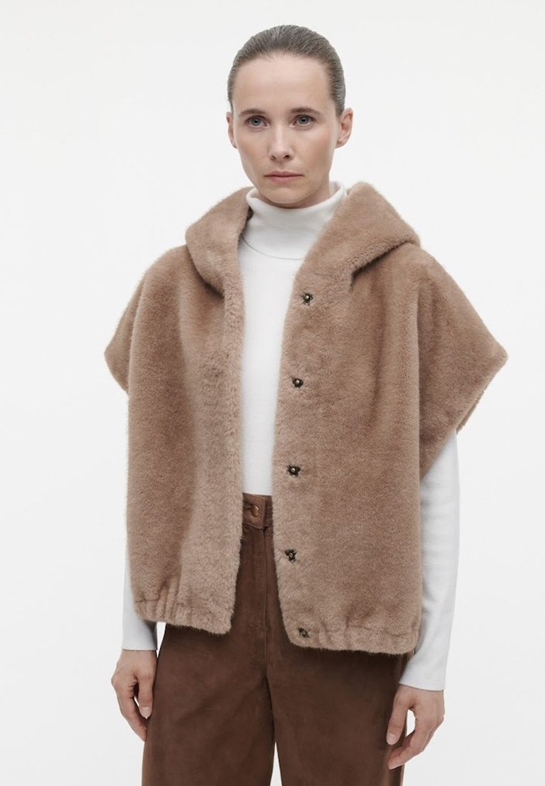 Faux fur kappe i lysebrun, med hætte, korte ærmer, frontknapper og blød tekstur. Båret over en hvid højhalset bluse og brune bukser.