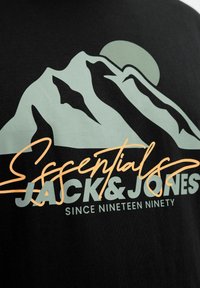 Sort t-shirt med et bjerggrafik i lys grøn og en orange kursiv tekst "Essentials" sammen med "JACK & JONES" og "SINCE NINETEEN NINETY."