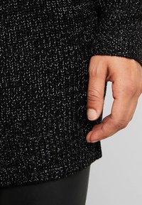 Musta neulepusero, jossa on teksturoitua lankaa ja hopeisia lankayksityiskohtia. Hihansuussa on resorineulos, ja käsi lepää helman päällä.