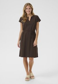 Robe brune à manches courtes avec un col en V et une ceinture nouée. Tissu lisse, longueur genoux, associée à des espadrilles beiges.