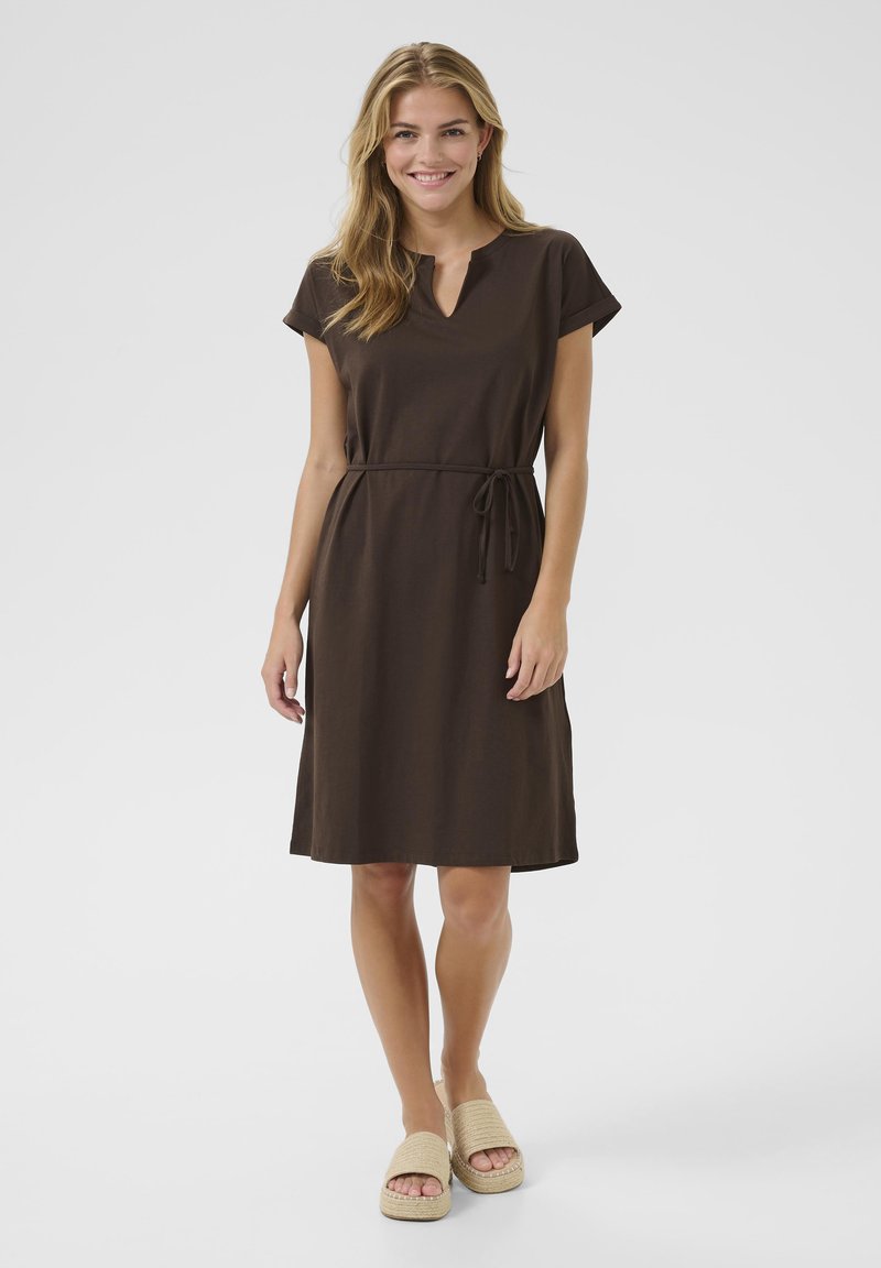 Robe brune à manches courtes avec un col en V et une ceinture nouée. Tissu lisse, longueur genoux, associée à des espadrilles beiges.