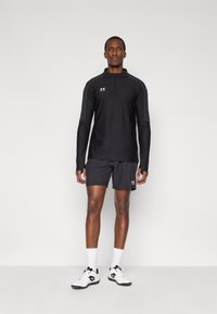 Μαύρο αθλητικό μπλουζάκι με μακρύ μανίκι και φερμουάρ μέχρι το στήθος, με γκρι λεπτομέρειες, σε συνδυασμό με μαύρο σορτς και λευκά αθλητικά παπούτσια με μαύρες λεπτομέρειες.