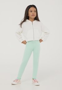 Felpa bianca con zip e tasche a marsupio, abbinata a leggings verde menta e sneakers con dettagli arcobaleno. Tessuto liscio, vestibilità casual.