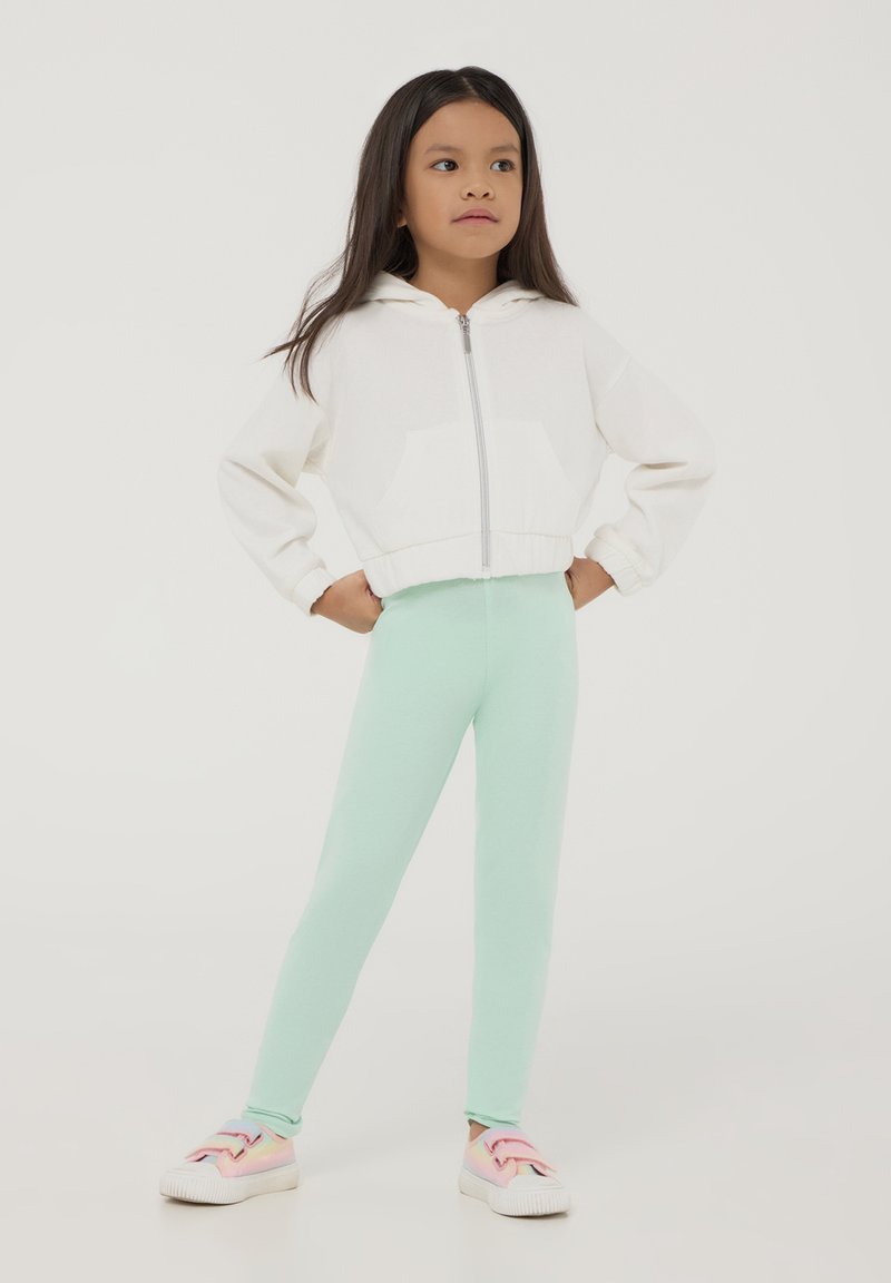 Felpa bianca con zip e tasche a marsupio, abbinata a leggings verde menta e sneakers con dettagli arcobaleno. Tessuto liscio, vestibilità casual.