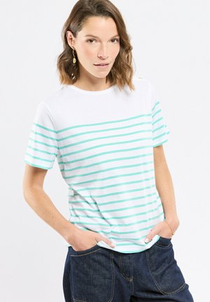 Femme aux cheveux bruns mi-longs portant des boucles d'oreilles dorées, un t-shirt blanc à rayures horizontales bleu clair, et un jean bleu foncé les mains dans les poches.