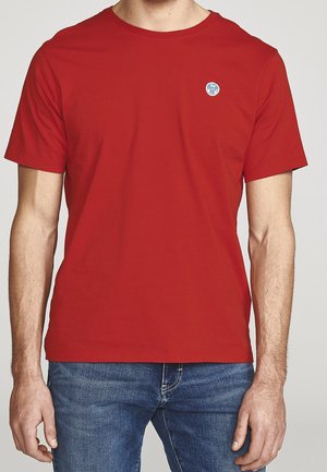 Homme portant un t-shirt rouge uni à manches courtes avec un petit logo rond bleu et blanc sur la poitrine gauche, associé à un jean en denim bleu.