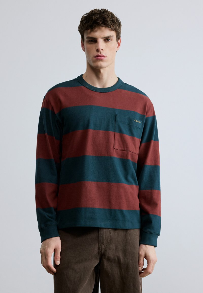 Μπλούζα με μακριά μανίκια, κόκκινες και τιρκουάζ οριζόντιες ρίγες, στρογγυλή λαιμόκοψη, και τσέπη στο αριστερό στήθος. Κατασκευασμένη από βαμβακερό ύφασμα.