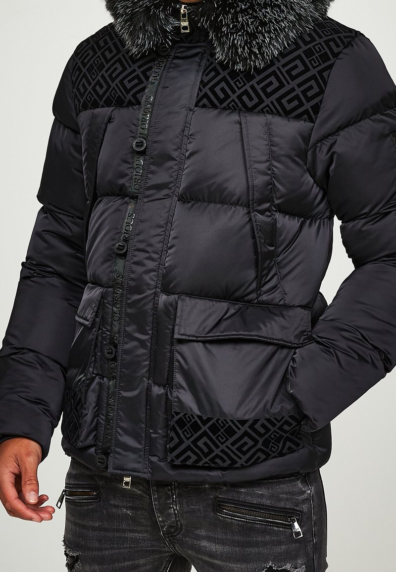 Sort quiltet pufferjakke med mønstrede paneler, frontlommer med klap, logo lynlås, og hætte med pelskant, båret sammen med slidsede sorte jeans.
