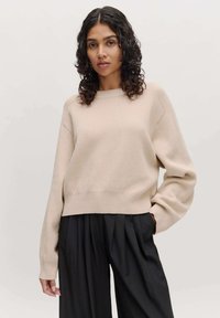 Beiger Pullover mit Rundhalsausschnitt, gerippten Bündchen und Saum. Weicher, leicht strukturierten Stoff. Kombiniert mit lockeren, schwarzen, plissierten Hosen.